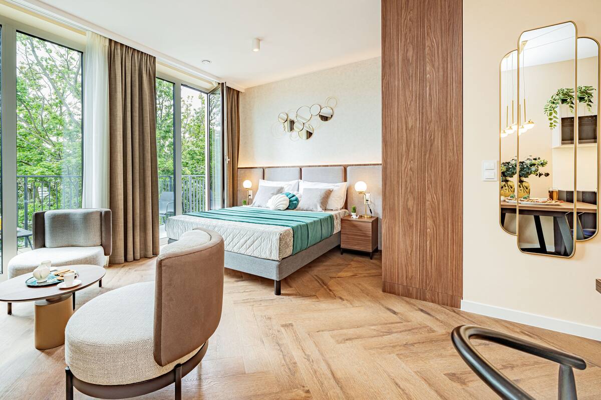Een moderne hotelkamer met een groot bed, een zithoek met een stoel en een tafeltje, en een balkon met uitzicht op bomen.