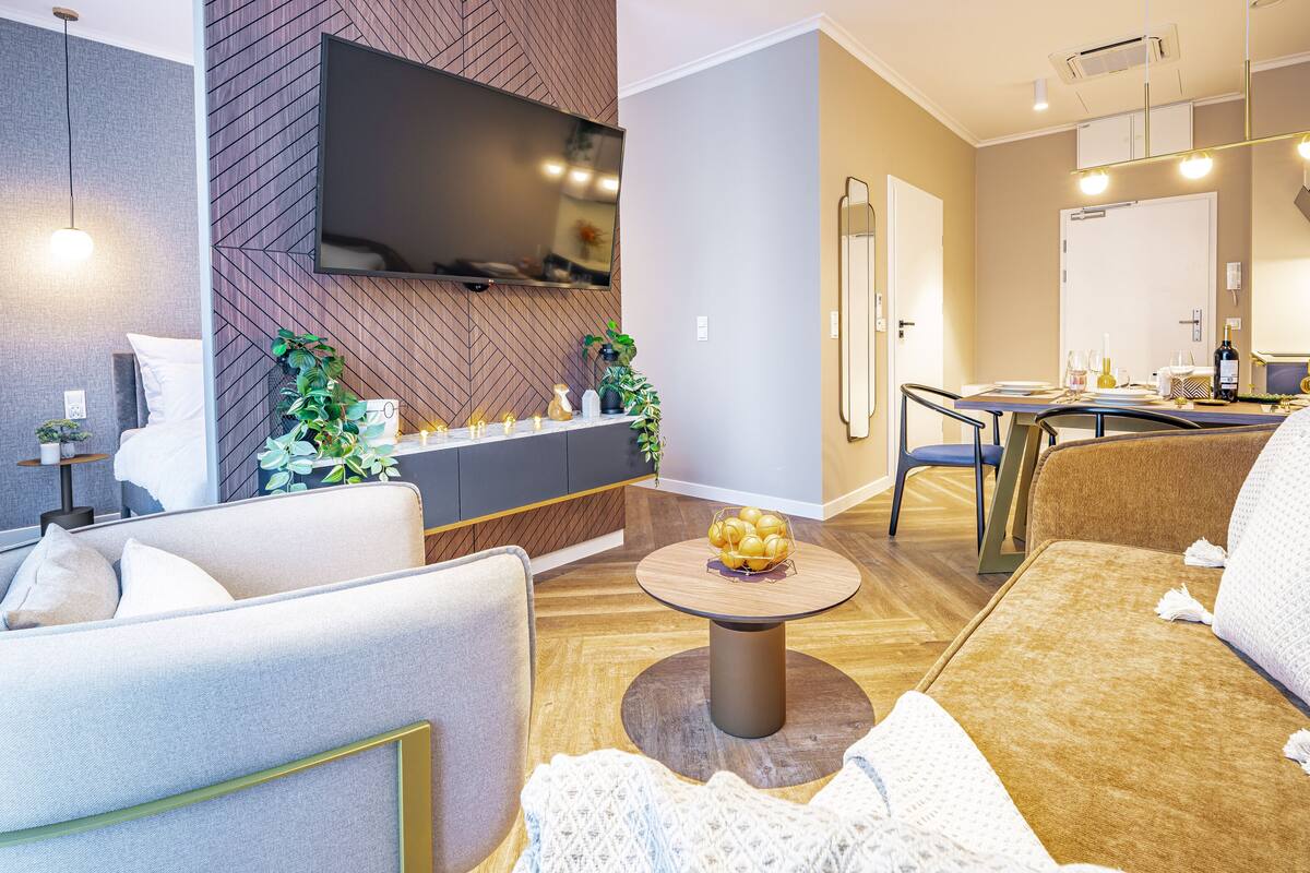 Een moderne woonkamer met een bank, fauteuil en salontafel. Er is een flatscreen televisie aan de muur bevestigd, een eettafel met stoelen en een keuken in de achtergrond zichtbaar.