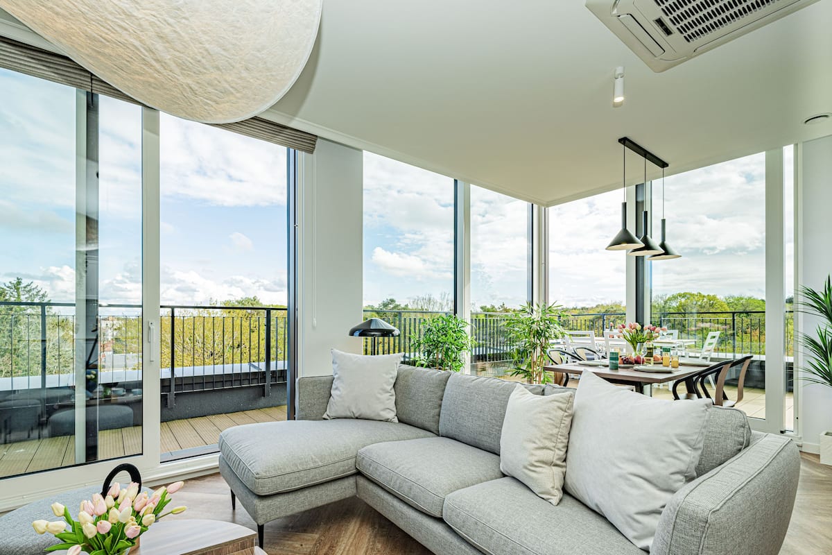 Een moderne woonkamer met een grote grijze hoekbank, een eethoek met een tafel en stoelen, en hoge ramen van de vloer tot het plafond met uitzicht op de omgeving.