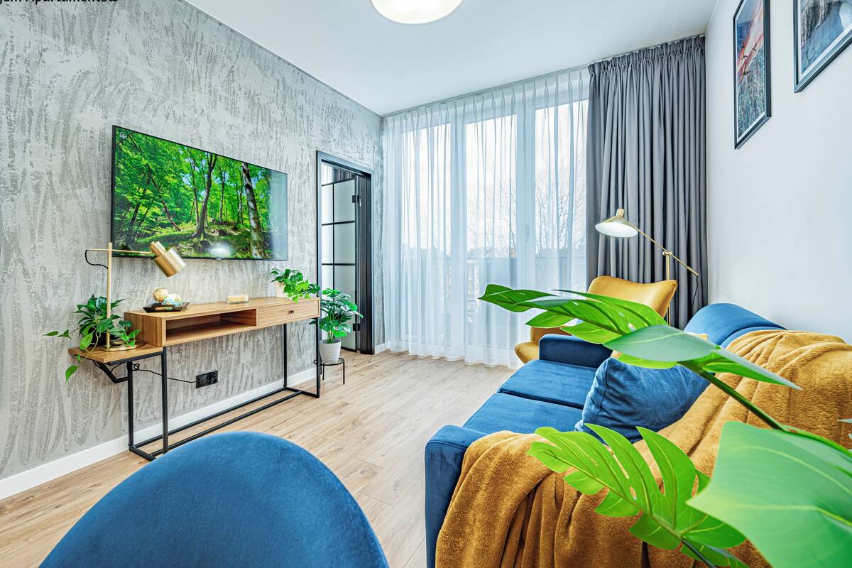 Een moderne woonkamer met een grote flatscreen televisie, een houten console met planten, een blauwe bank en een groot raam met gordijnen.
