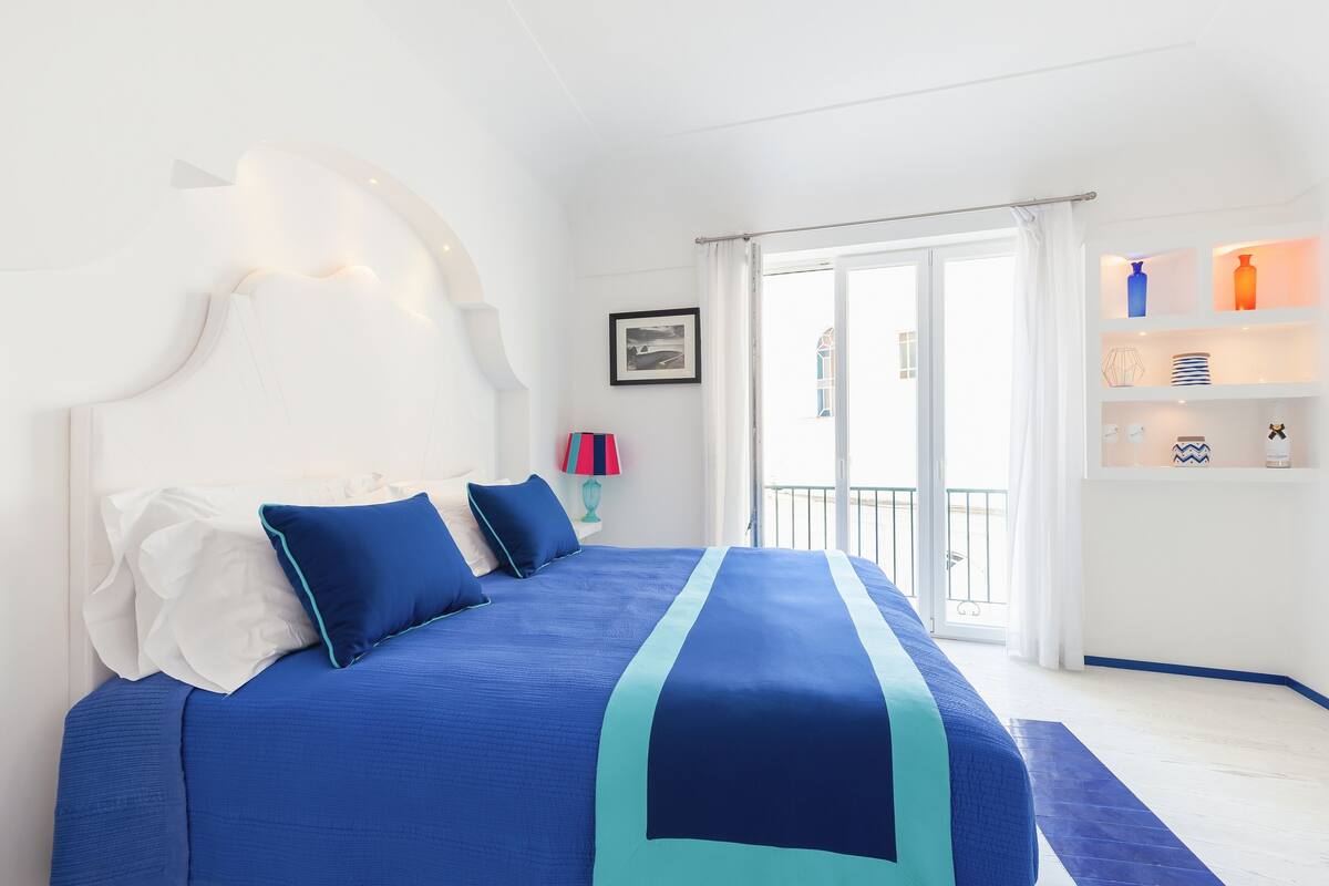 A bedroom with a large bed, white walls, a balcony with a railing, and a shelf with decorative items.