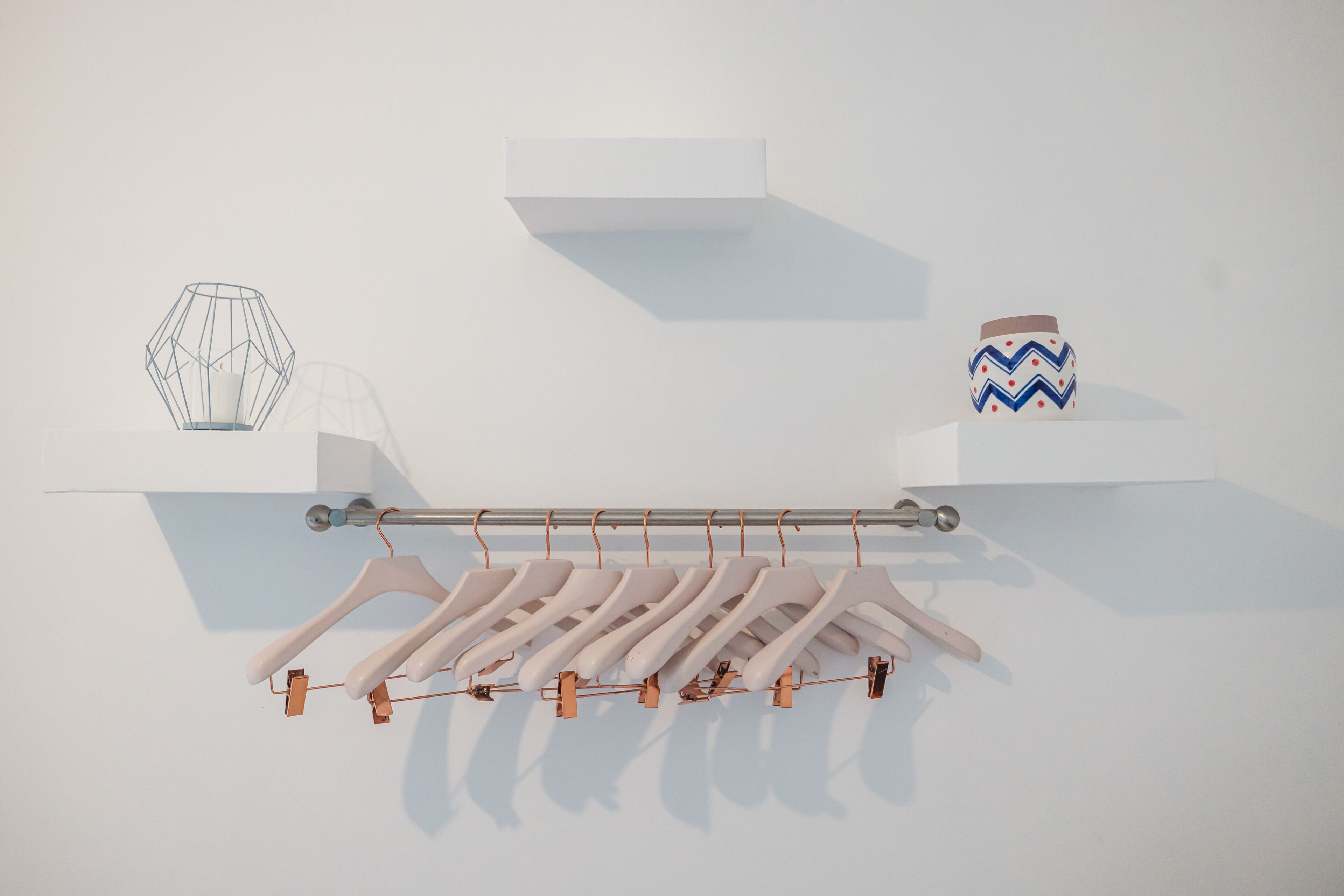 Een minimalistische kamer met een witte muur, een metalen rek met witte kledinghangers en een geometrische vaas op een plank.