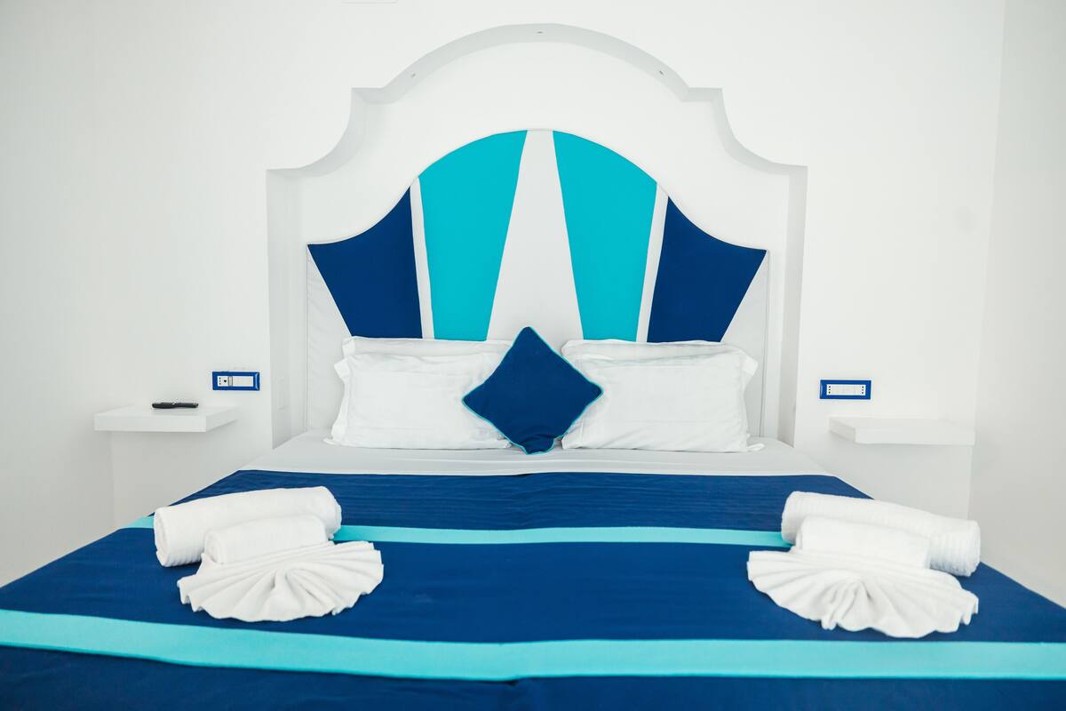 A hotel room with a large bed, white headboard, and blue bedding. There are two bedside tables with lamps and a power outlet on each.