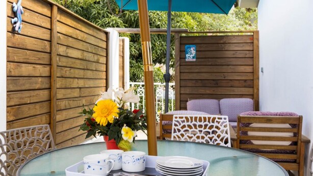 A round glass table with a tray of white dishes, a vase of yellow flowers, and a wooden fence in the background. - Appartementmaïthé 3 (Saint-Paul)