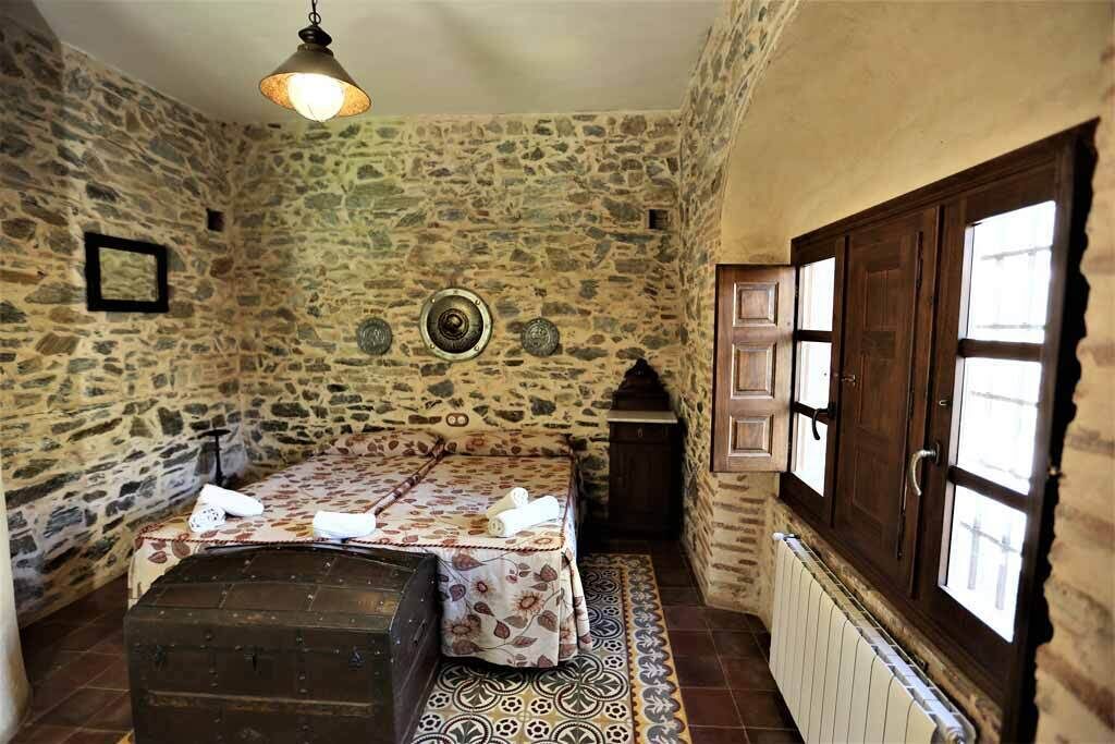 A stone-walled room with a bed, a chest, a small table, and a window with wooden shutters.