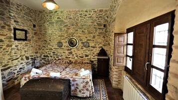 A stone-walled room with a bed, a chest, a small table, and a window with wooden shutters.