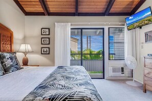 A bedroom with a bed, a nightstand, a lamp, a fan, and a TV on the wall.