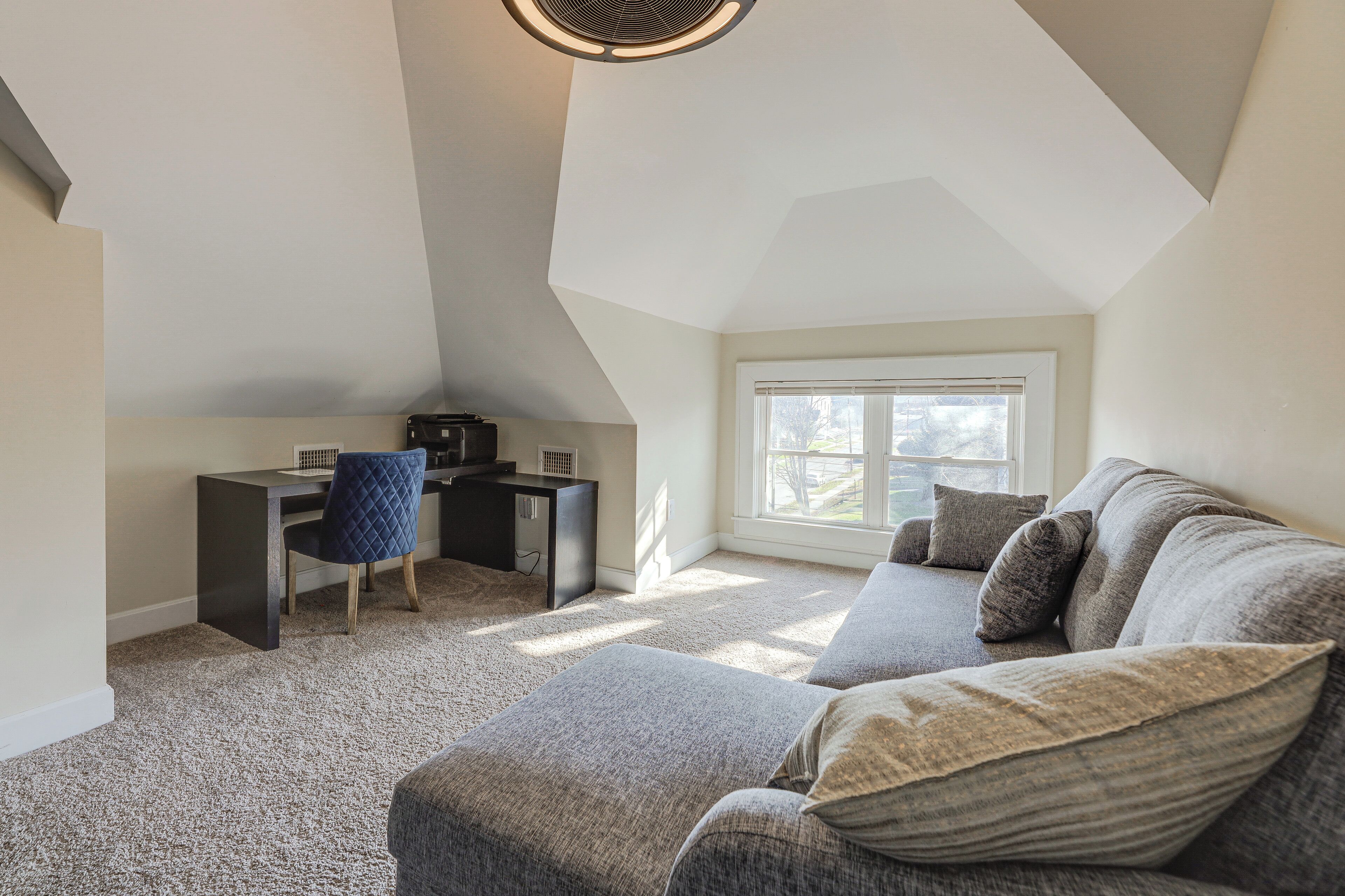 A loft-style bedroom with a sloped ceiling, a desk with a chair, a gray sofa, and a large window providing natural light.