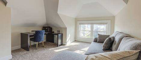 A loft-style bedroom with a sloped ceiling, a desk with a chair, a gray sofa, and a large window providing natural light.