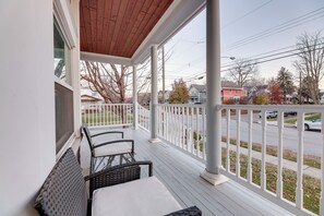 Un porche cubierto con dos sillones de mimbre y una mesa, con vista a una calle con casas y automóviles.