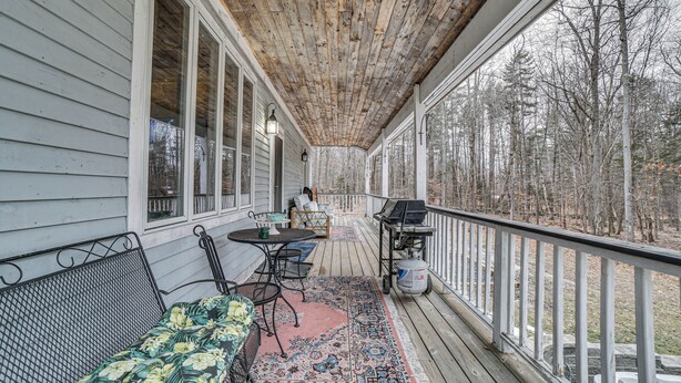 A covered porch with a wooden ceiling, a dining table with chairs, a grill, and a view of trees.