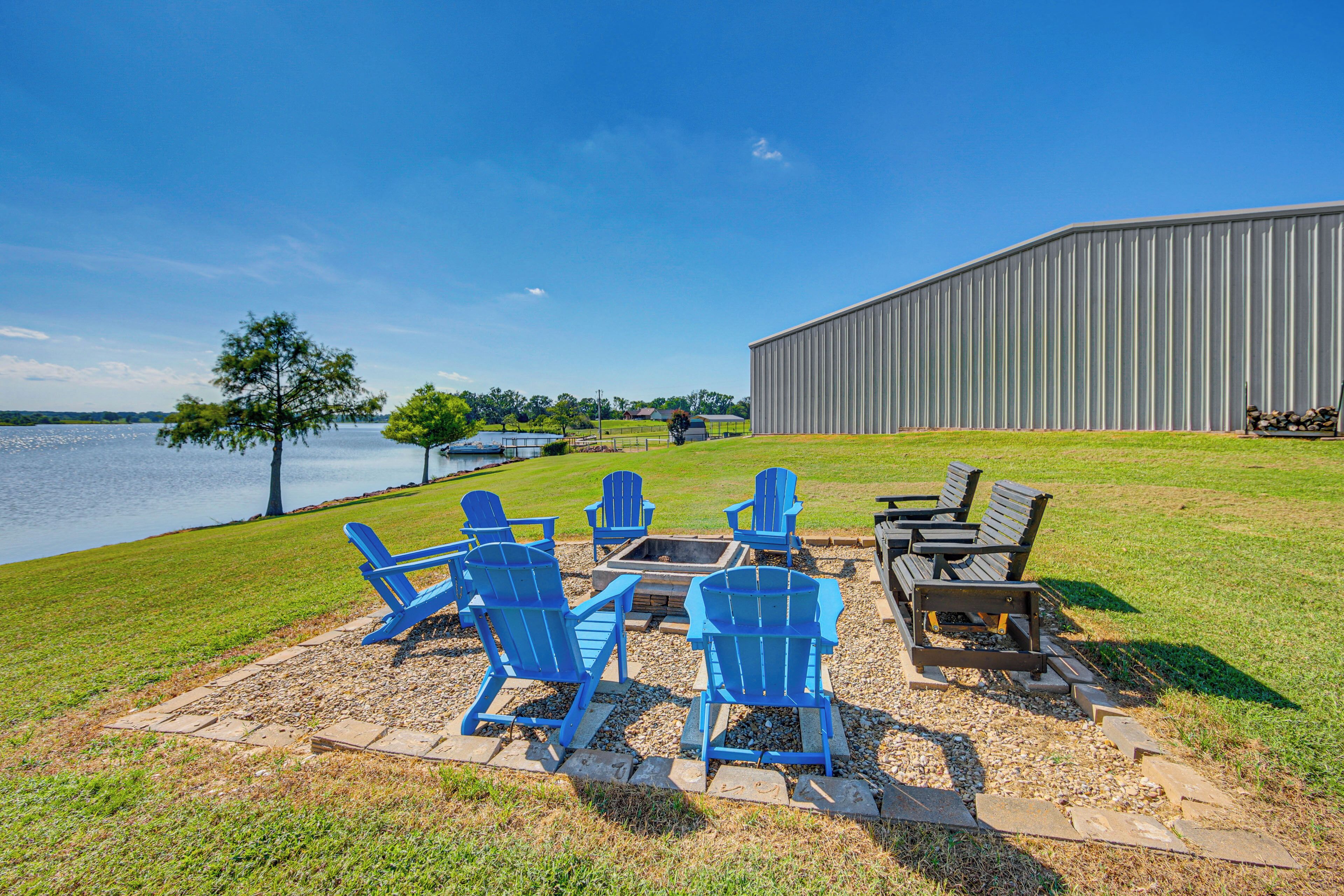 A lakeside patio with blue Adirondack chairs, a fire pit, and a large metal building in the background.