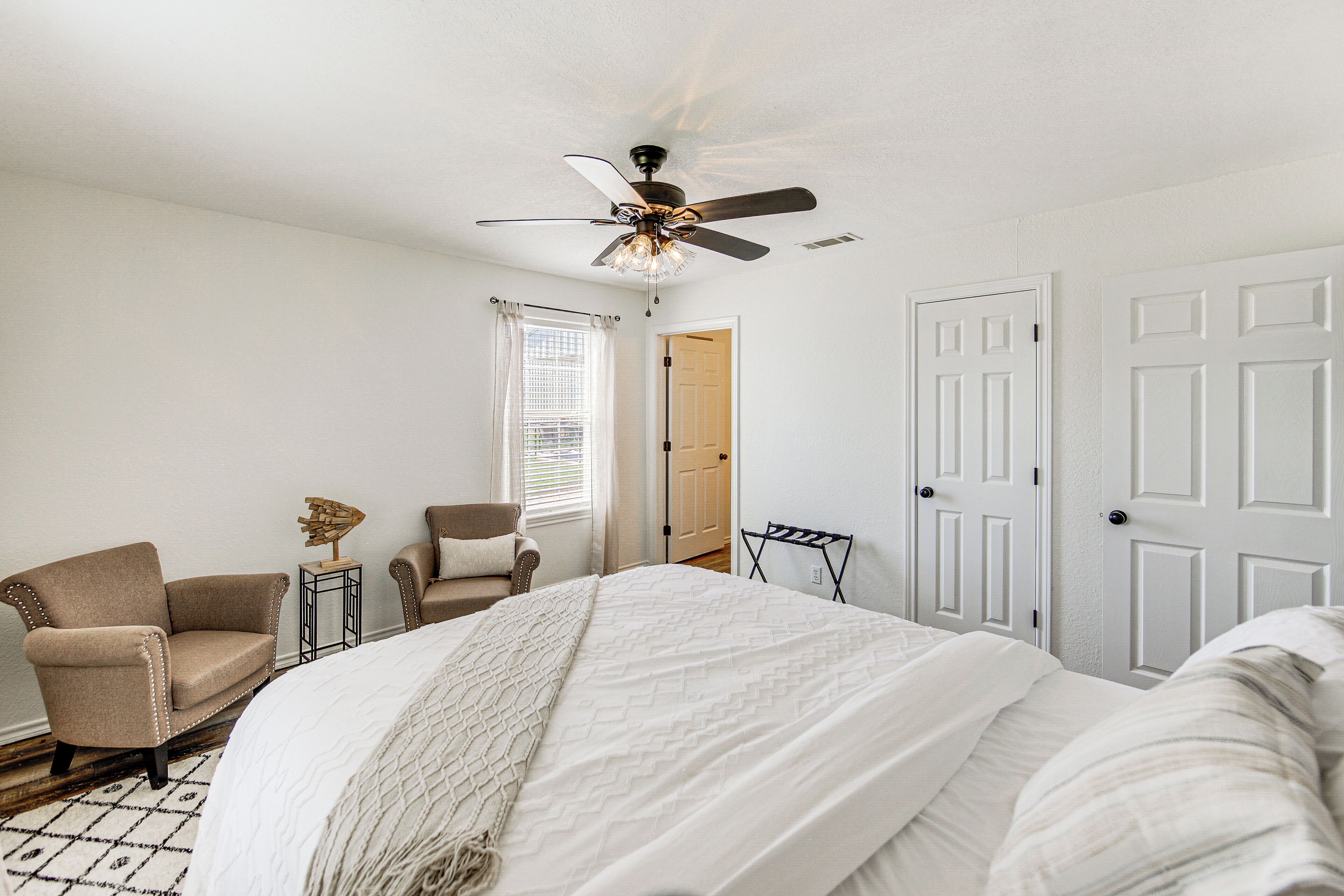 A bedroom with a bed, two chairs, a ceiling fan, and a door.