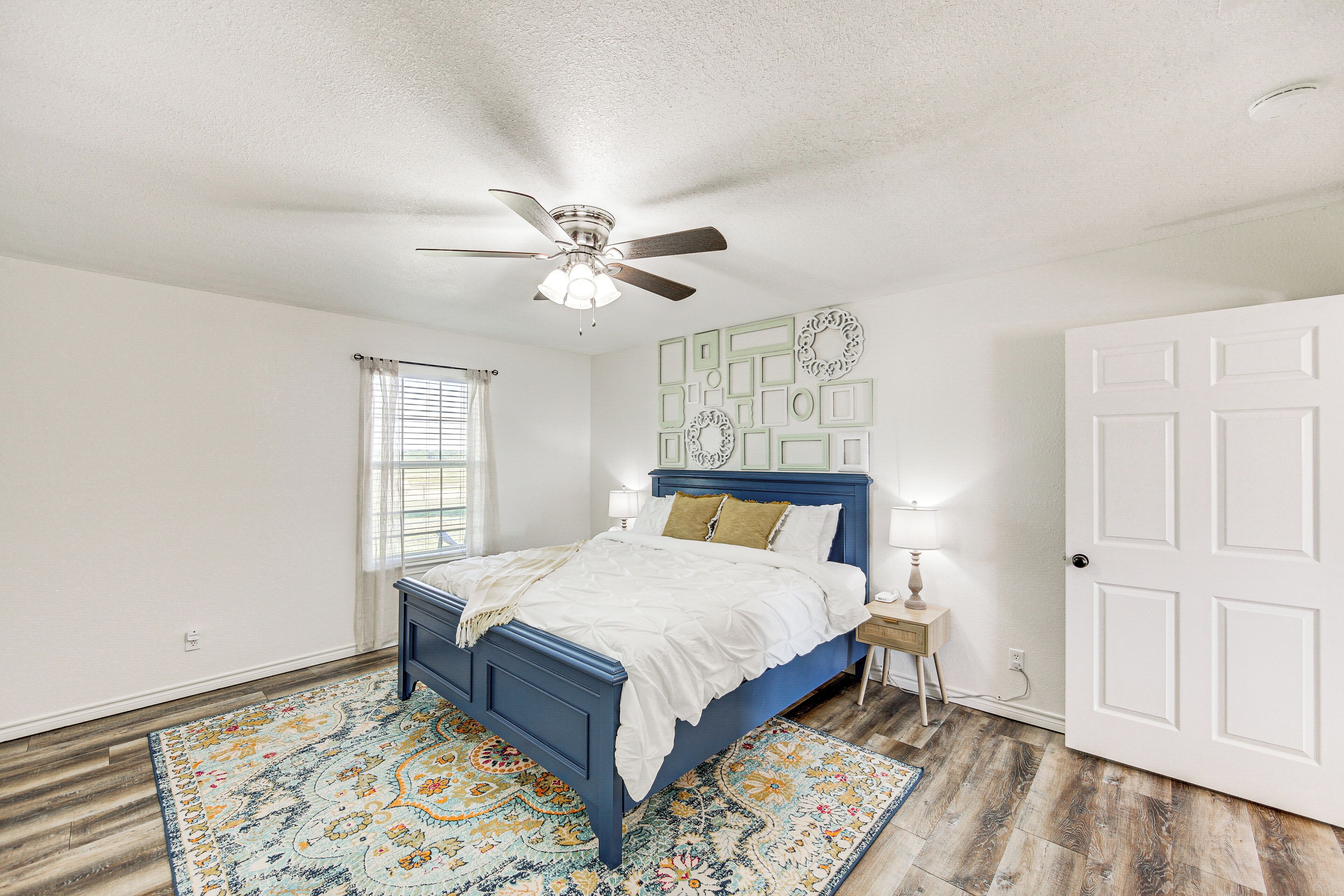 A bedroom with a bed, a ceiling fan, a window with blinds, a door, and a patterned rug.