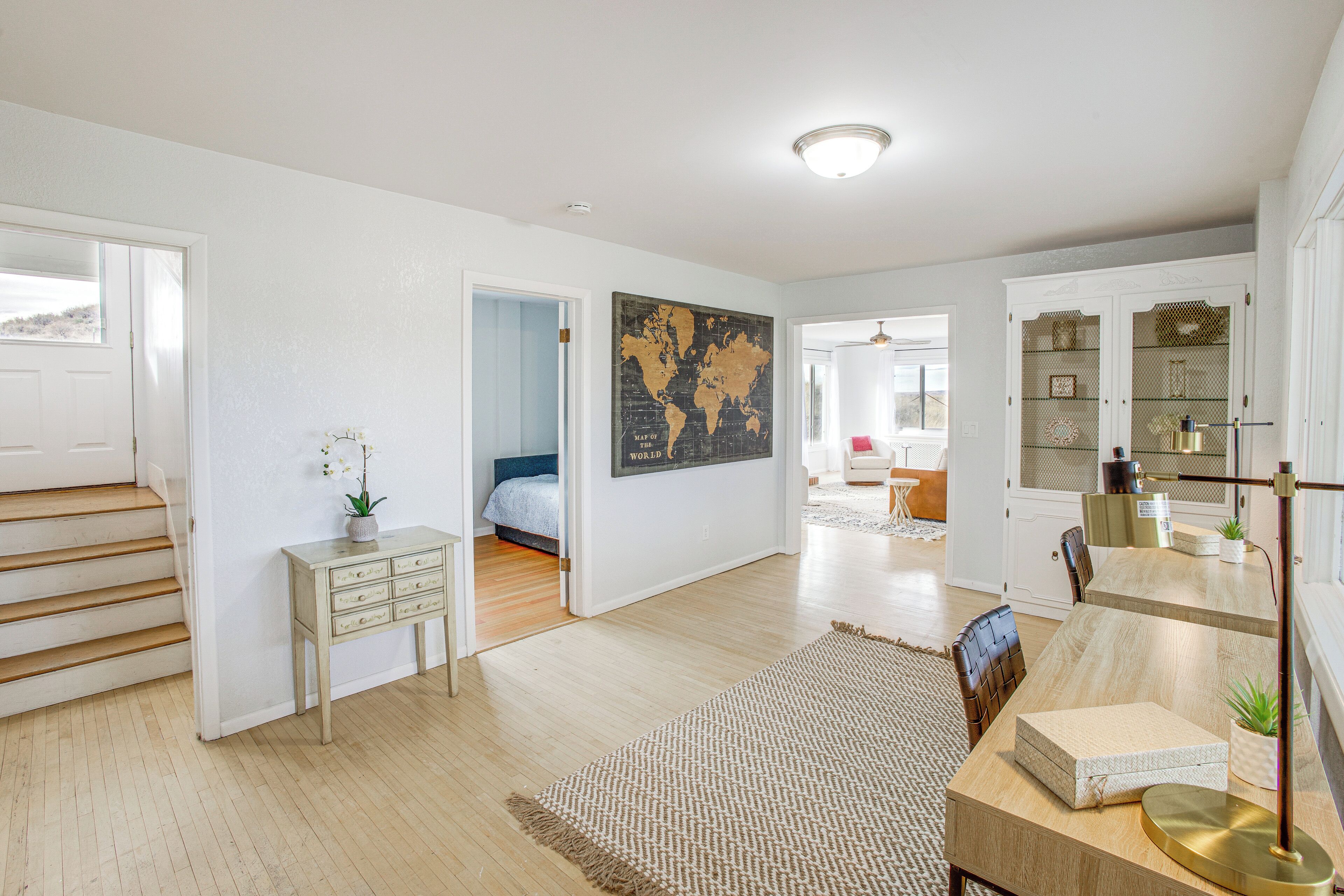 A spacious living area with a wooden floor, a world map on the wall, a desk with drawers, a bed in the adjacent room, and a dining area with a table and chairs.