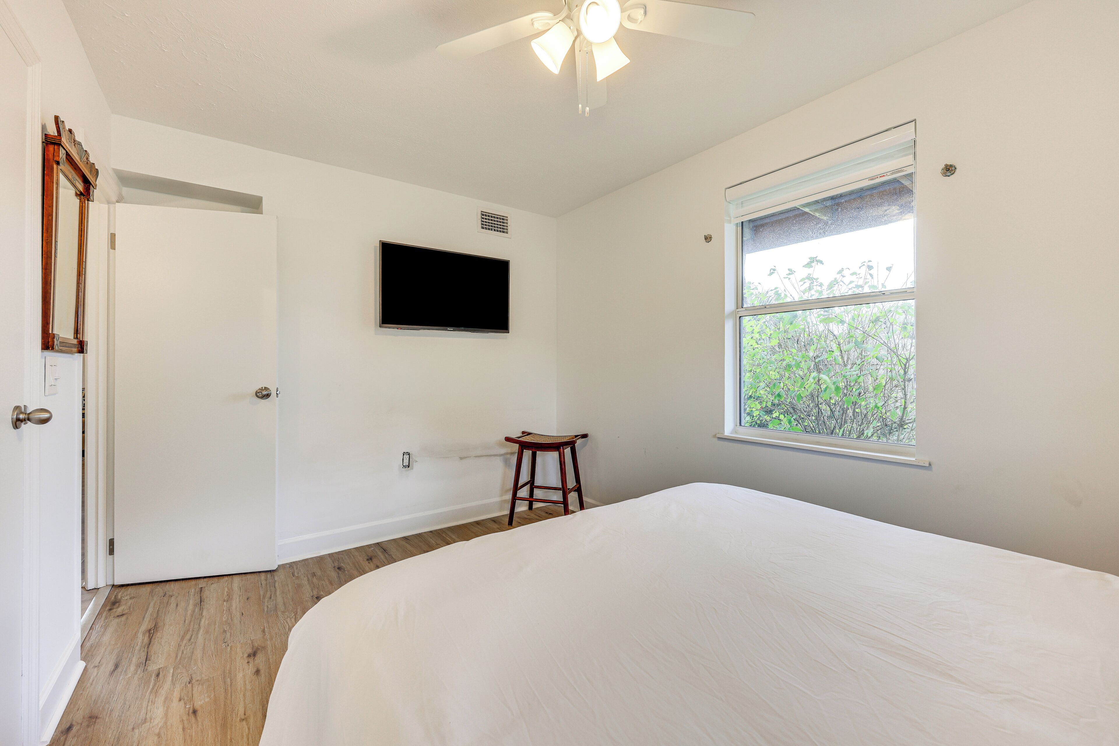A bedroom with a large bed, a wooden floor, a ceiling fan, a mounted TV, a window with a view of greenery, and a door.