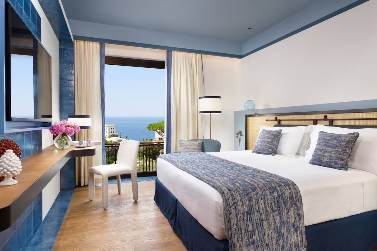 A hotel room with a large bed, a desk with a vase of flowers, a chair, and a view of the sea.
