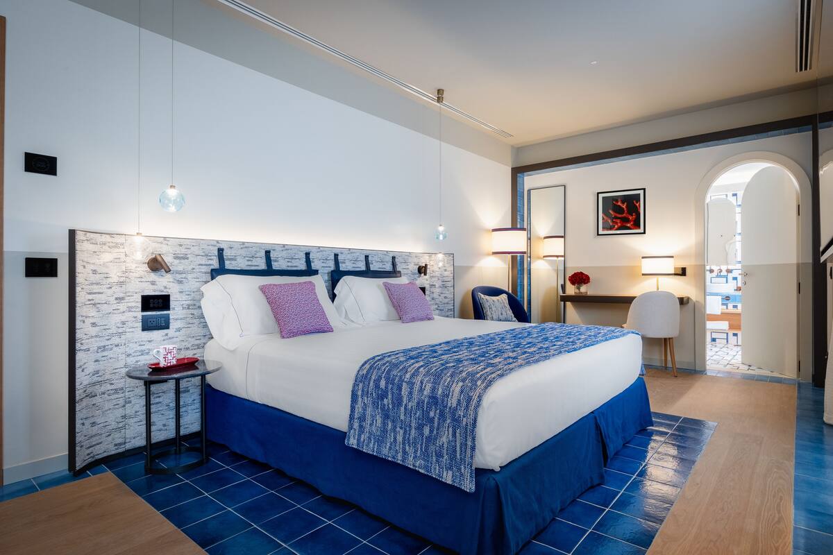 A modern hotel room with a large bed, a small side table, a desk with a chair, and a bathroom visible through an archway.