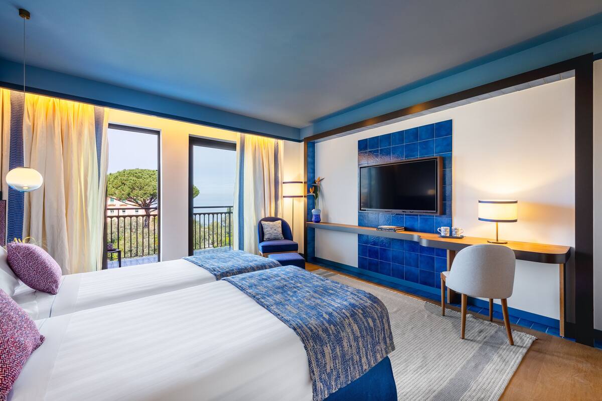 A hotel room with two beds, a TV, a desk, and a balcony with curtains.