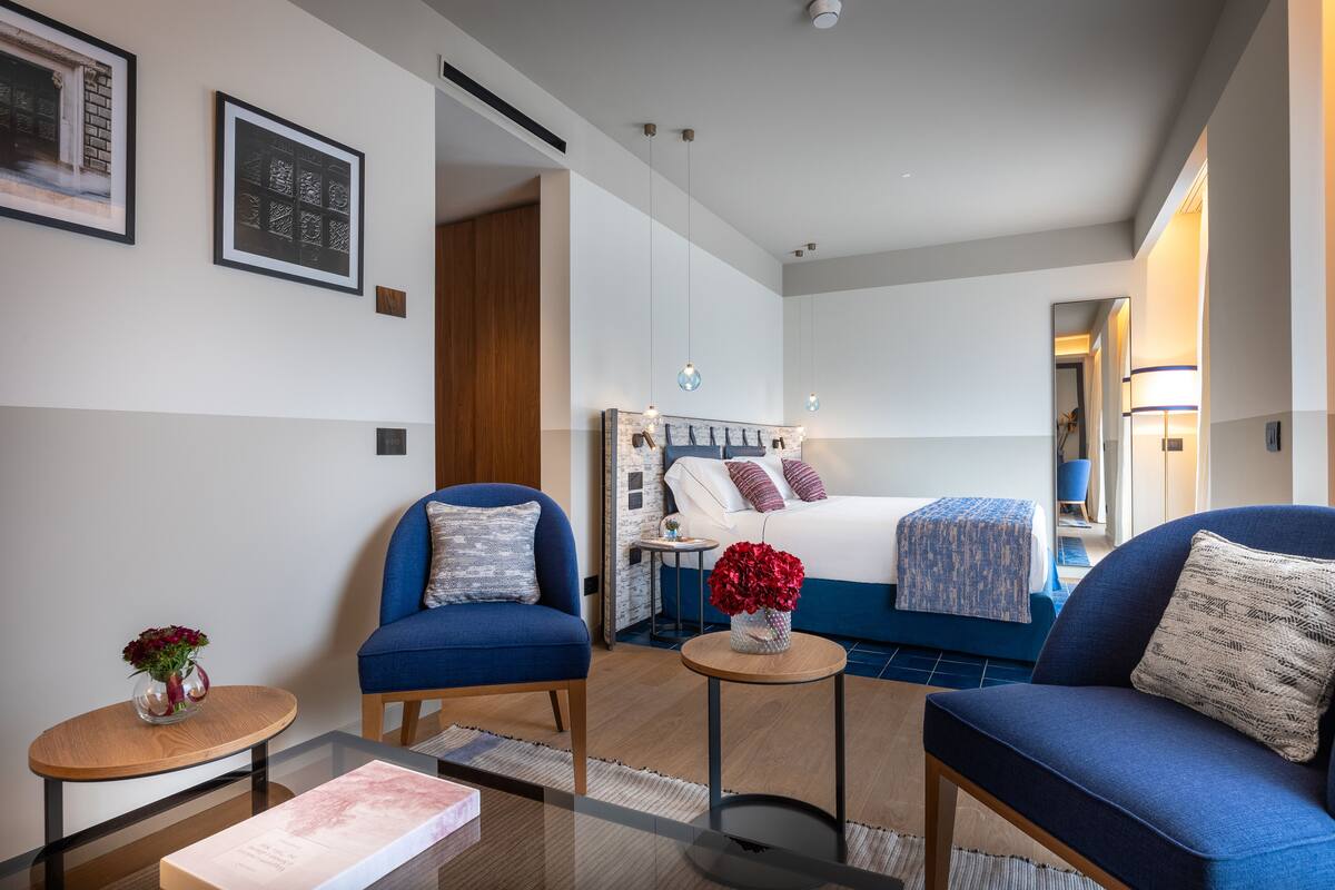 A modern hotel room with a large bed, two blue armchairs, a small table with a vase of flowers, and a mirror on the wall.