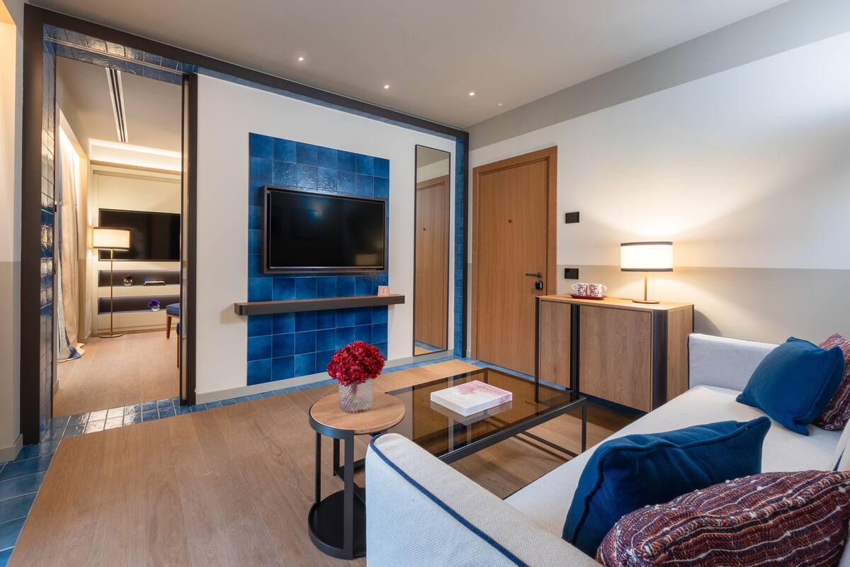 A modern hotel room with a glass coffee table, a sofa, a TV mounted on a blue-tiled wall, and a wooden door.