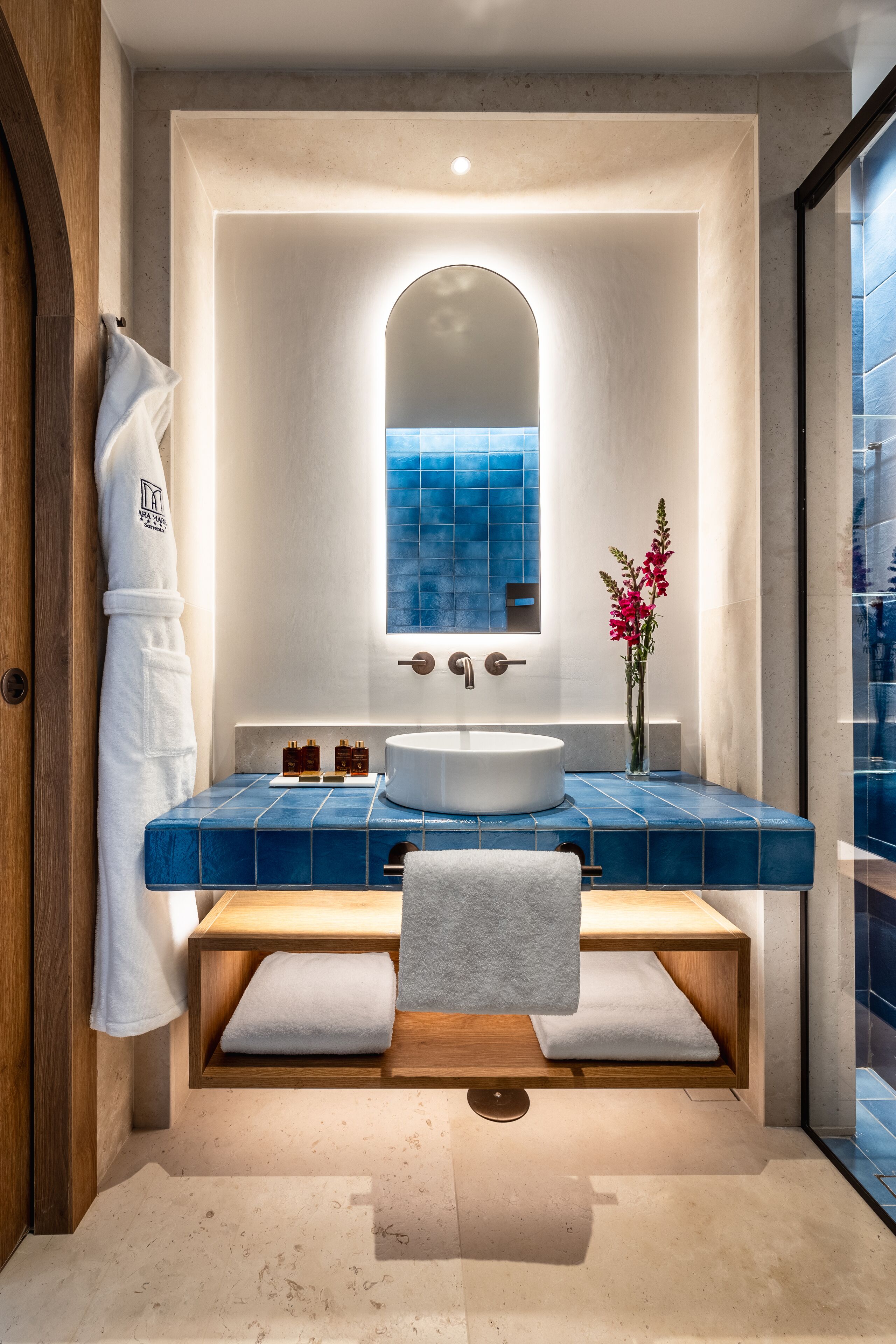 Un bagno moderno con uno specchio grande, un lavabo bianco, piastrelle blu, una panca in legno e un accappatoio bianco appeso al muro.