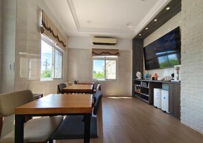 Un comedor con mesas y sillas de madera, un televisor de pantalla plana montado en una pared de ladrillo y una zona de cocina con electrodomésticos.