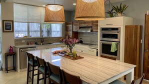 A modern kitchen with a large island, two hanging wicker pendant lights, a built-in oven, and a window with a view of the outdoors.