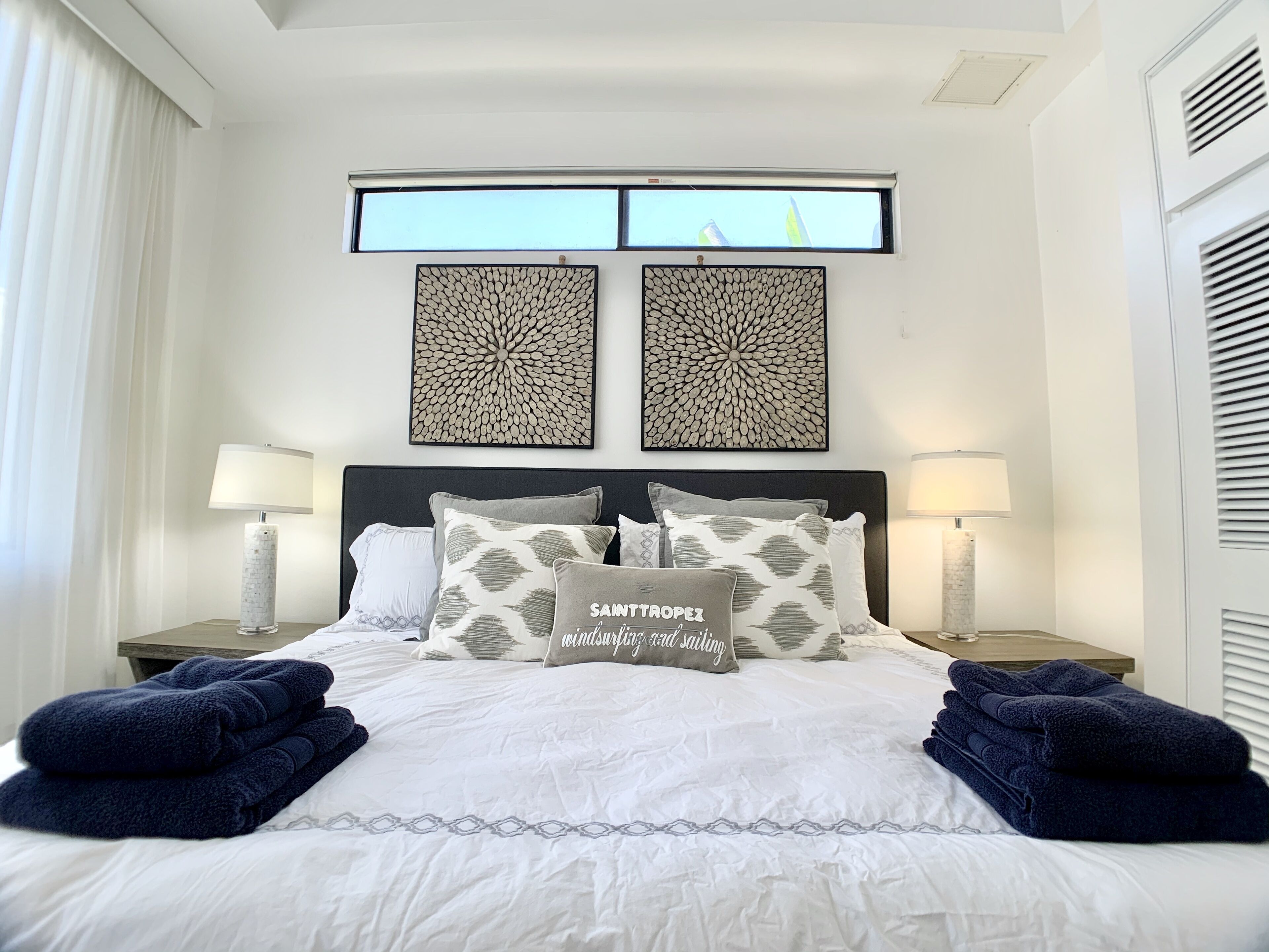 A neatly made bed with decorative pillows, two wall-mounted artworks, and a window with a view of flags.