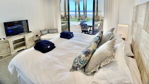 A hotel room with a large bed, a TV, a view of the beach, and a balcony with outdoor furniture.