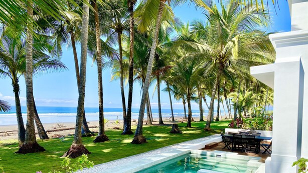 A pool area with a view of the beach and ocean, surrounded by palm trees and a white building.
