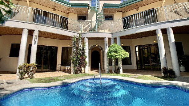 A two-story building with a central courtyard featuring a swimming pool, surrounded by columns and potted plants. - Villa Arenas: Private Pool & Easy Beach Access in Jaco by RedAwning (Jacó)