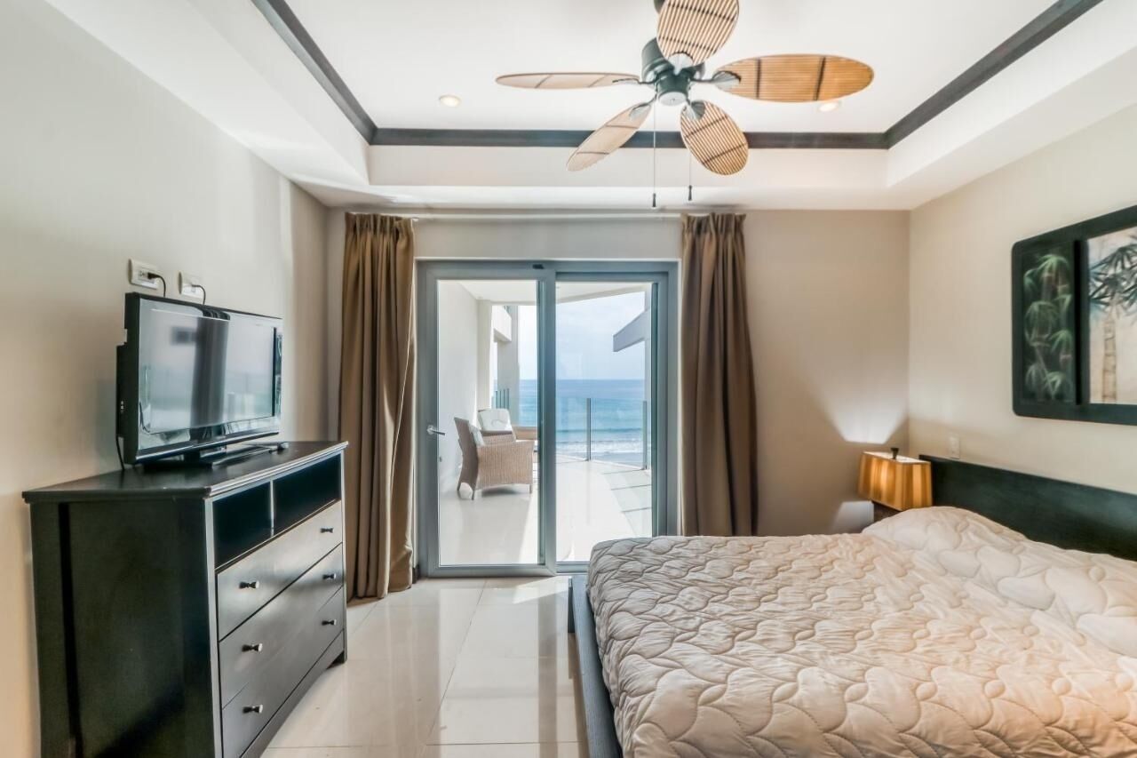 A hotel room with a bed, a television on a dresser, a ceiling fan, and a view of the ocean through a sliding door.