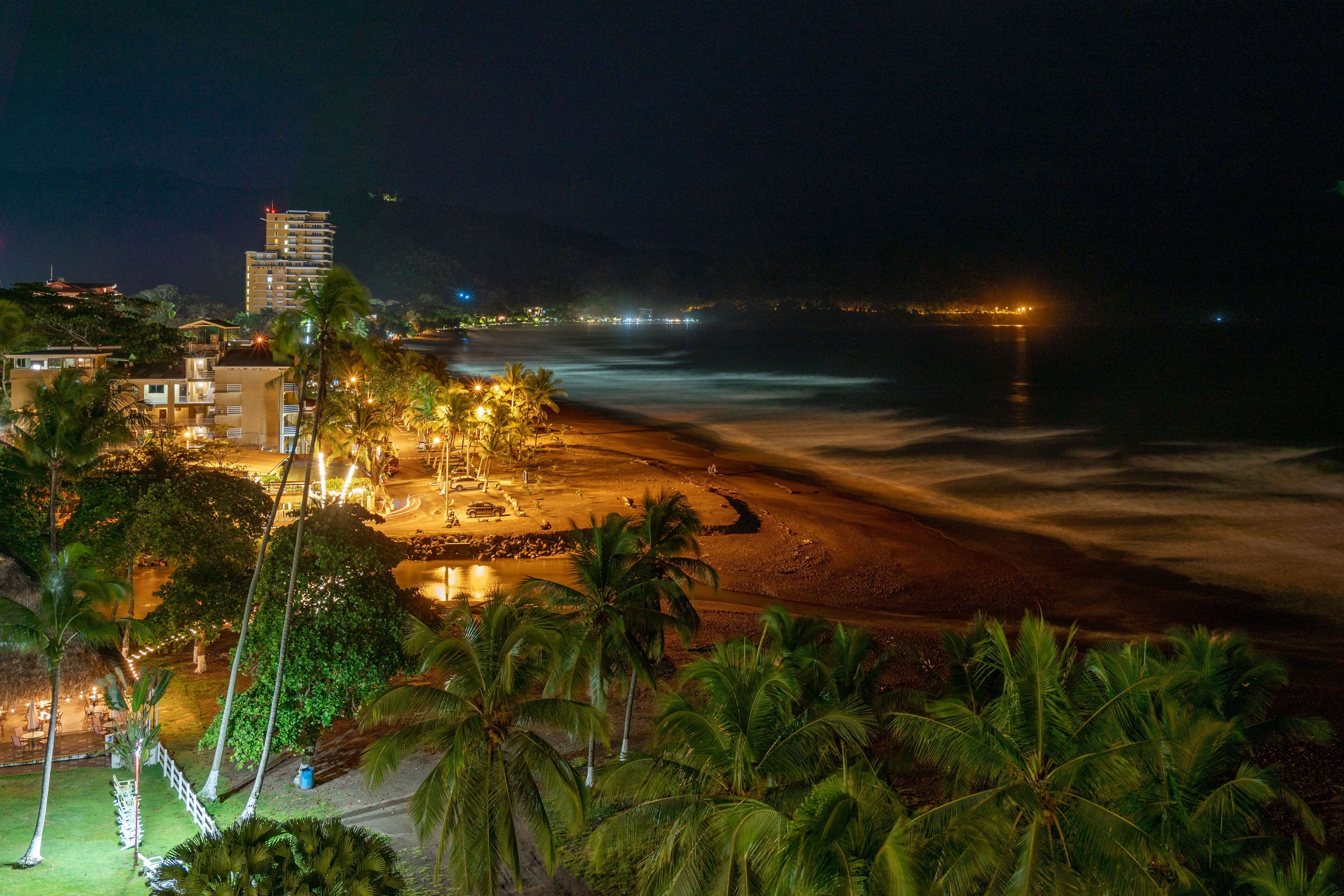 Vista de la playa con un hotel, palmeras y un paisaje urbano lejano de noche.