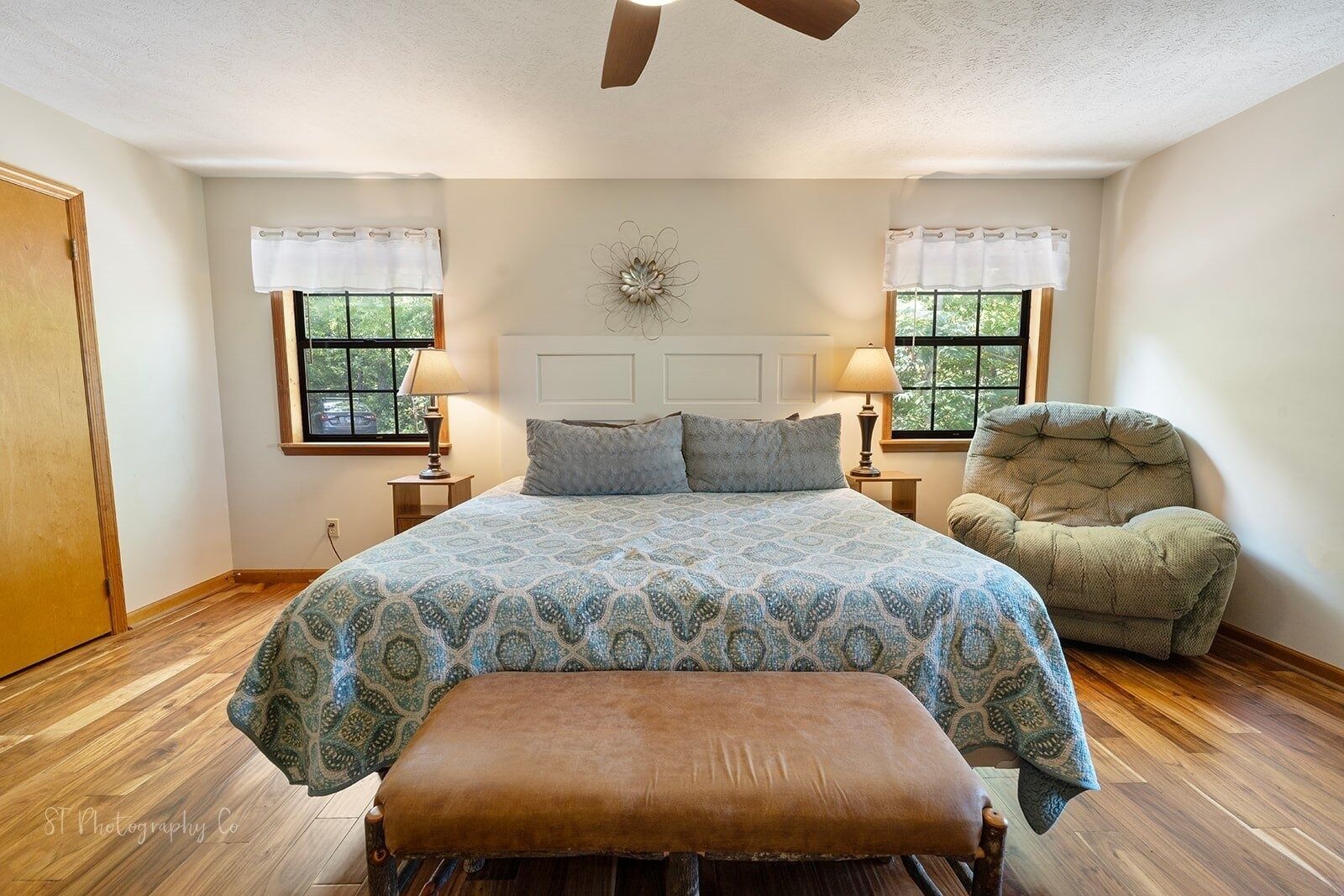 A bedroom with a large bed, two bedside tables, a chair, a bench, and a ceiling fan.