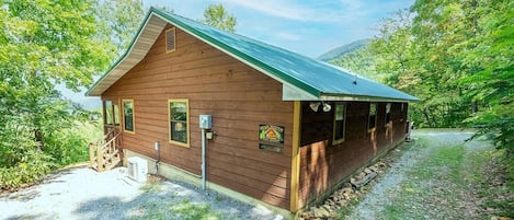 Una cabaña de madera con techo metálico, rodeada de árboles y un sendero de grava.