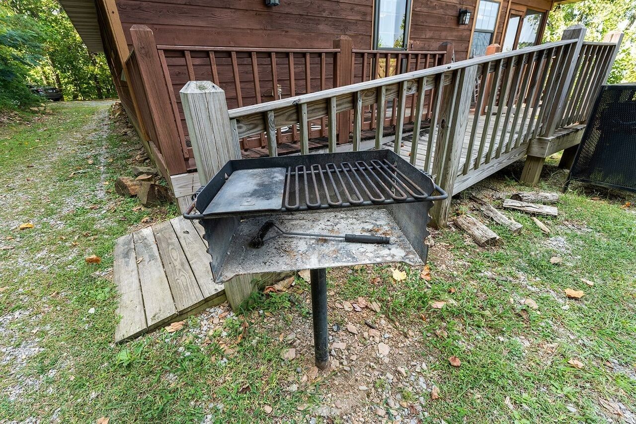 An outdoor grill with a metal grate and a brush, set on a wooden platform in a grassy area with a wooden deck and a house in the background.