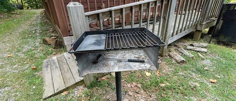 Een buitenbarbecue met een metalen rooster en een borstel, geplaatst op een houten platform in een grasrijke omgeving met een houten terras en een huis op de achtergrond.