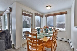 A dining room with a wooden table set for four, surrounded by chairs, with a view of a snowy landscape through large windows. - Fall Line Condo A5 1 Bedroom Condo (Killington)