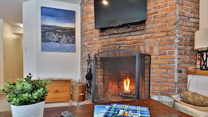 A living room with a brick fireplace, a mounted TV, a potted plant, and a magazine on the table. - Fall Line Condo A5 1 Bedroom Condo (Killington)