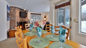 A dining area with a brick fireplace, wooden chairs, and a table set with green placemats and vases. - Fall Line Condo A5 1 Bedroom Condo (Killington)