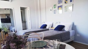A modern bedroom with two beds, a dining area with a table set for two, and a kitchen visible through an open door. - Apartamento Céntrico y Luminoso (San Nicolas)