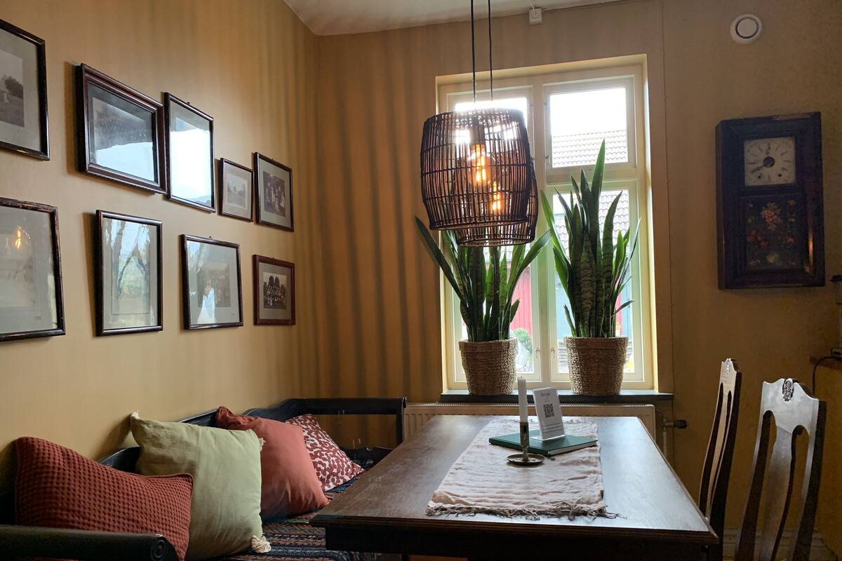 A dining area with a wooden table, chairs, a sofa with cushions, framed pictures on the wall, potted plants by the window, and a hanging light fixture.