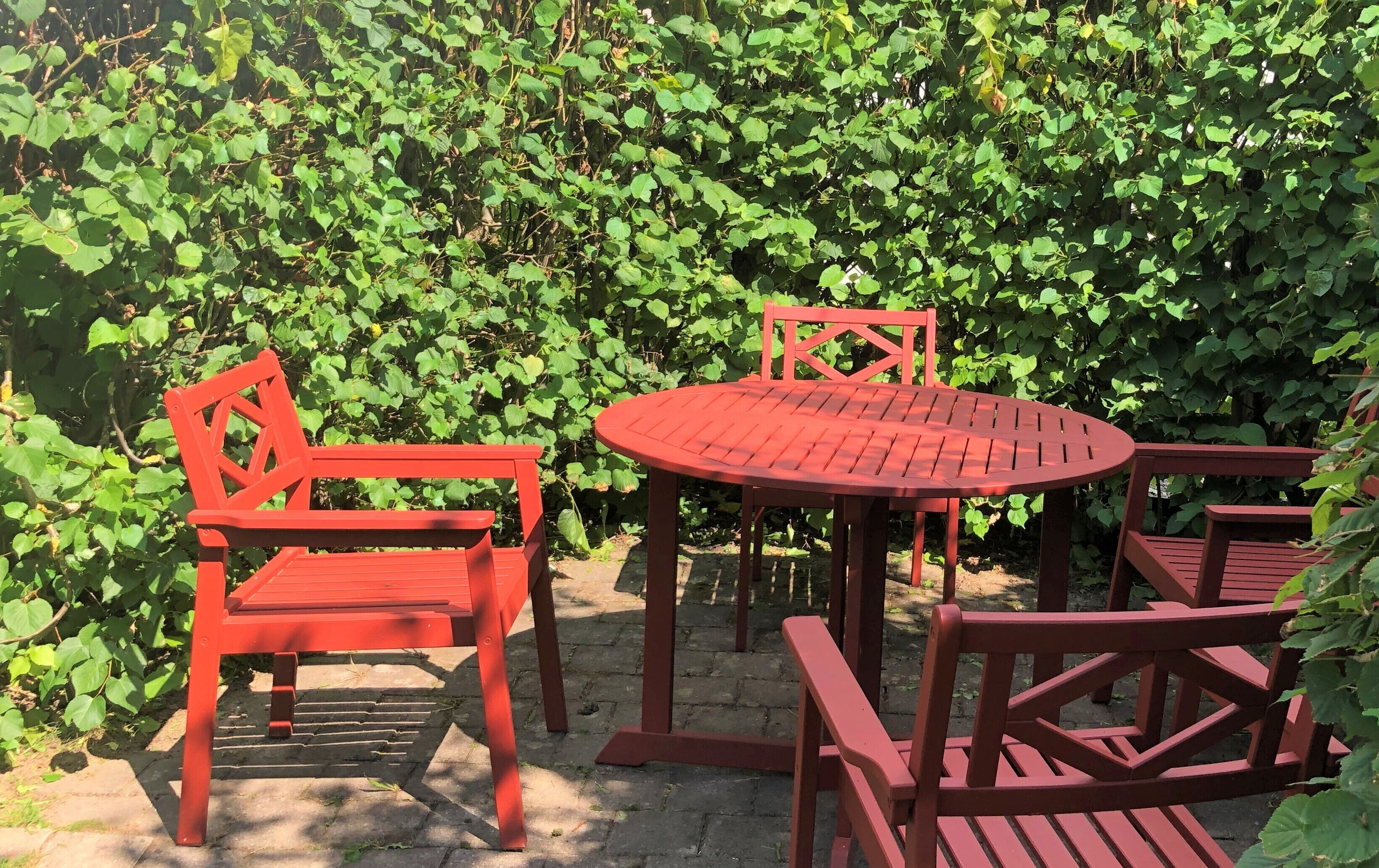 A red metal table and chairs set outdoors with a dense green hedge in the background.