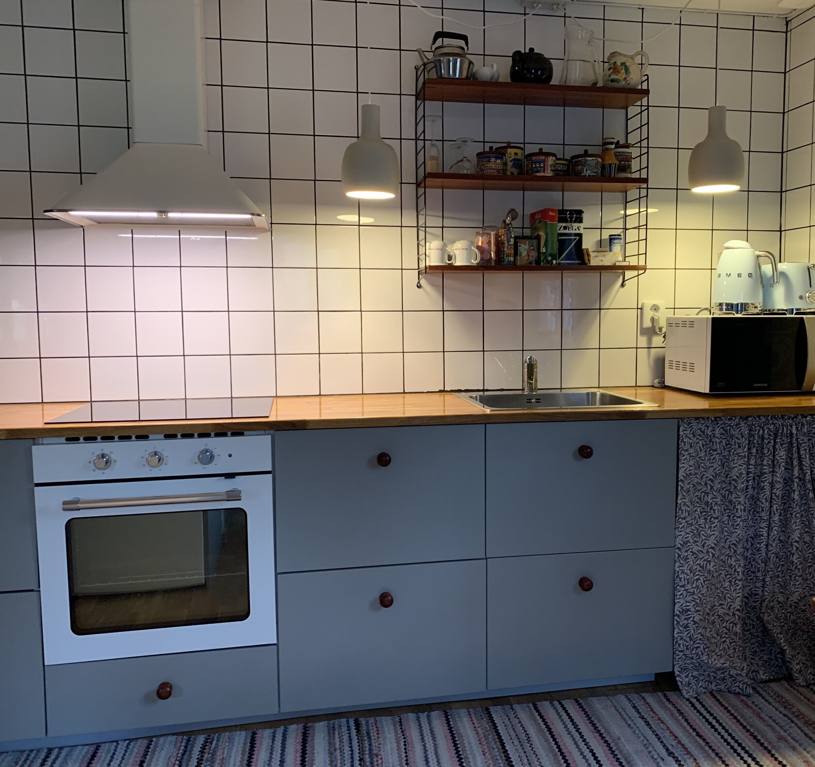 Cocina compacta con horno empotrado, lavamanos y cafetera sobre la encimera. Hay armarios de madera y un tapete estampado en el suelo.