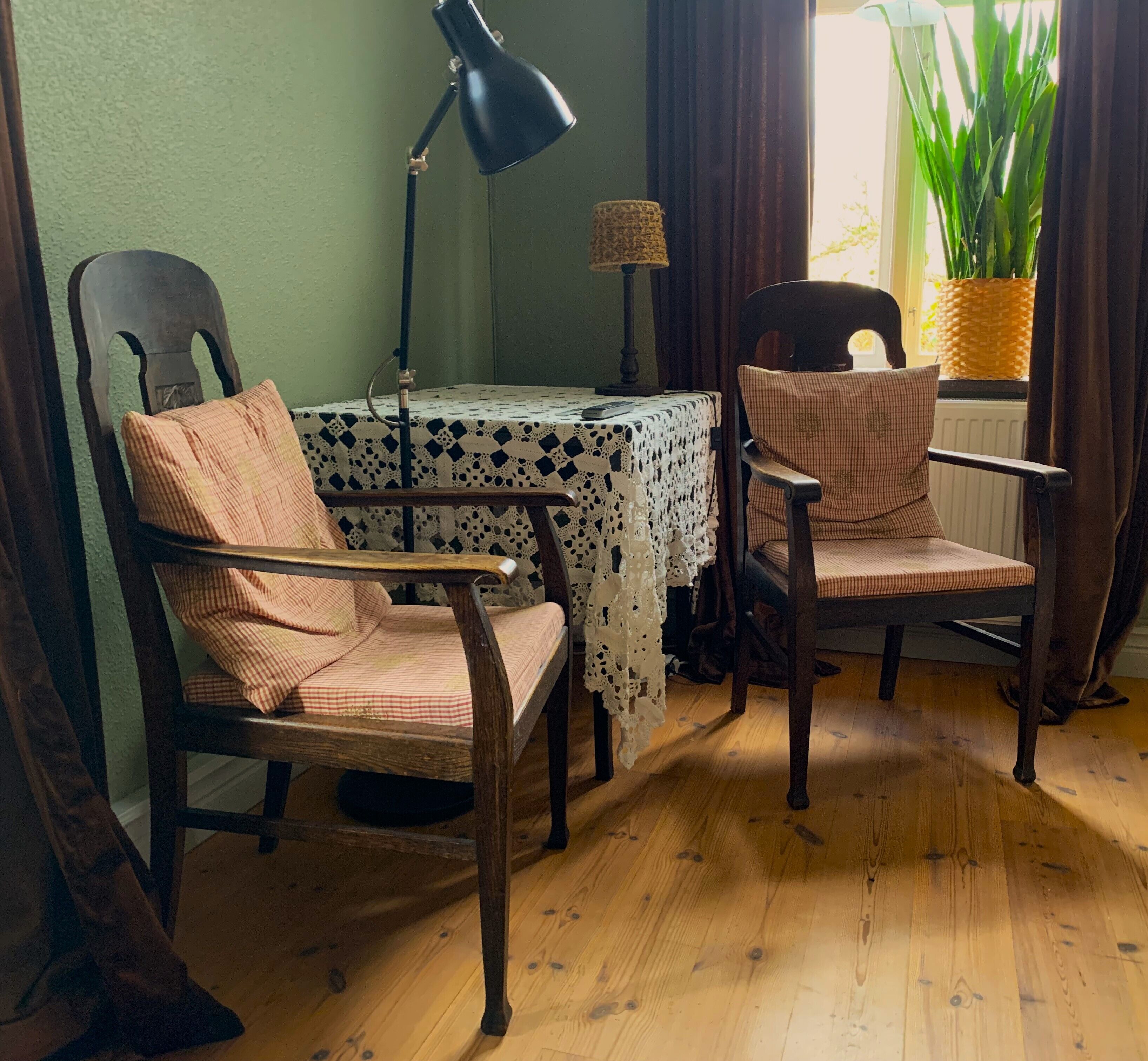 A room with two wooden chairs, a table with a lace tablecloth, a black floor lamp, and a potted plant by the window.