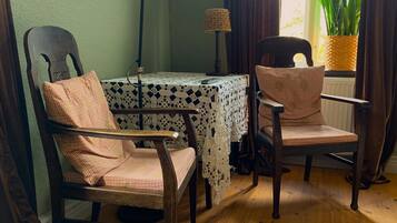 A room with two wooden chairs, a table with a lace tablecloth, a black floor lamp, and a potted plant by the window.