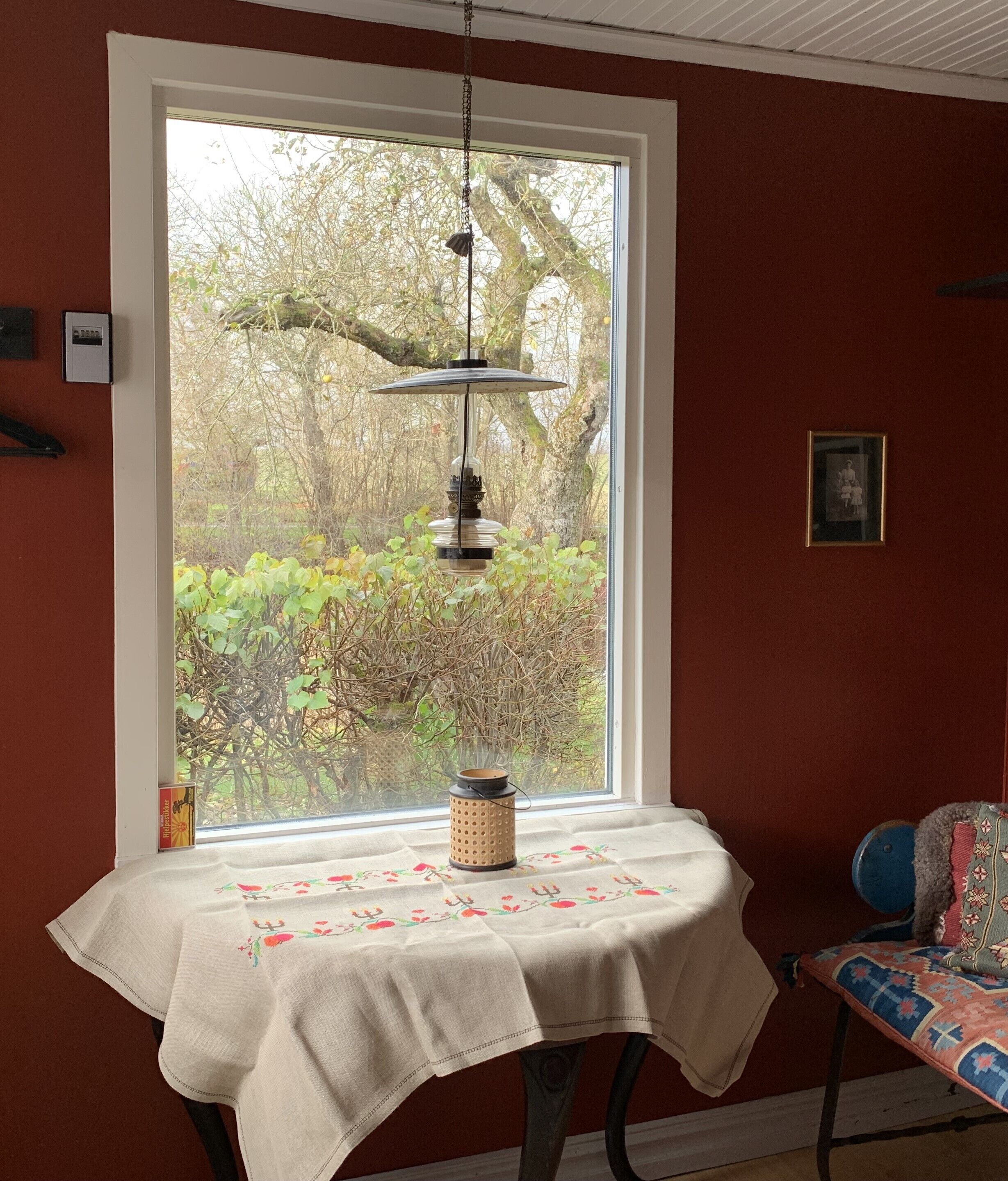 Una habitación con un ventanal grande, una mesa con mantel floral, un farol colgante y una silla con cojín estampado.