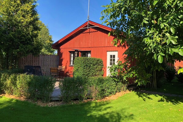 Un edificio de madera roja con puerta y ventanas blancas, rodeado de follaje y una valla de madera.