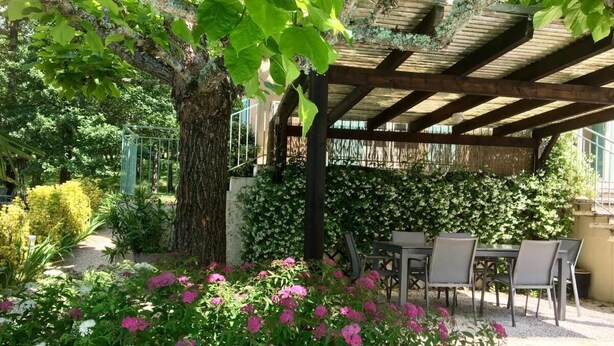 A covered outdoor seating area with a pergola, surrounded by lush greenery and flowering plants. - LES FEUILLANTINES (Saint-Cyprien)